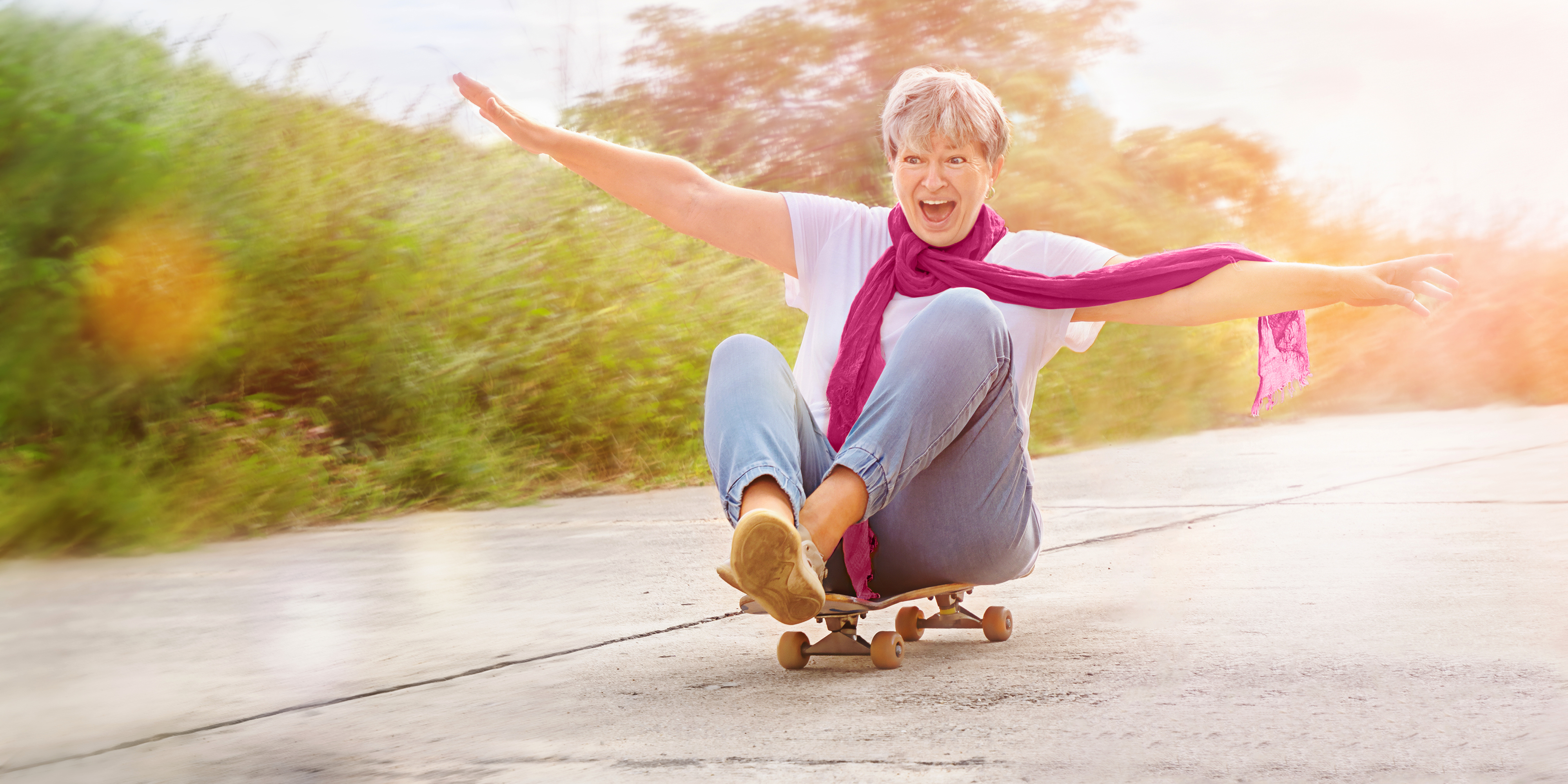 Ältere Frau auf einem Skateboard, Arme ausgebreitet, lächelnd. Blaue Jeans, weißes T-Shirt, pinker Schal.